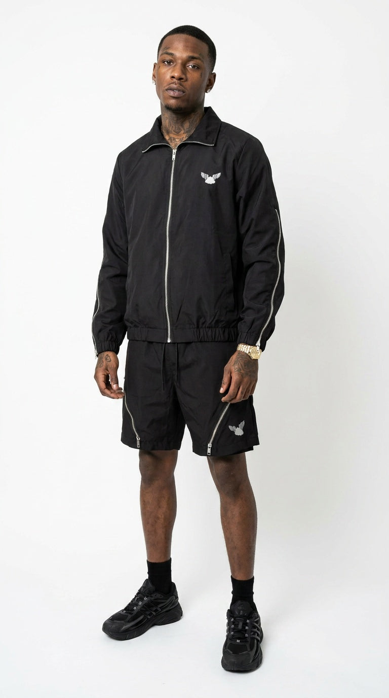 Full-body front studio shot of a male model wearing the coordinated iLLionaire stealth black lightweight track jacket and matching shorts set, highlighting the prominent silver zippers down the sleeves and across the thighs.