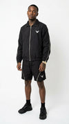 Full-body front studio shot of a male model wearing the coordinated iLLionaire stealth black lightweight track jacket and matching shorts set, highlighting the prominent silver zippers down the sleeves and across the thighs.